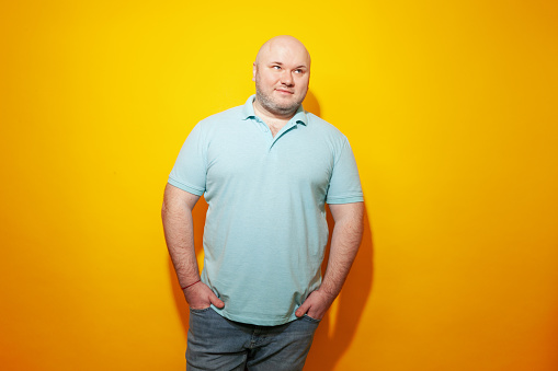 Studio portrait of overweight mid adult white man with shaved head, wearing blue polo shirt, against a yellow background
