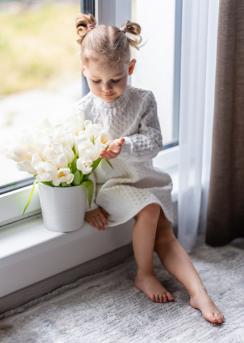 Little girl sitting by window with tulip flowers bouquet. Happy child, indoors. Mother's day, valentine's day or birthday concept. High quality photo