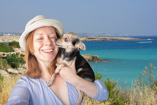 traveling with dogs, happy tourist in a hat takes a selfie with a small puppy in his arms against the background of the sea bay, a cute pet kisses the owner, travel snapshot