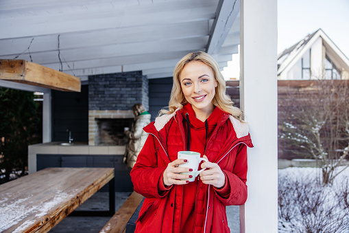 Cheerful cute young Caucasian blonde woman with a coffee mug leaning against the gazebo post