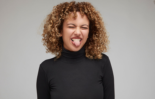 Young woman with curly hair closing eyes and sticking out tongue in front of white background.