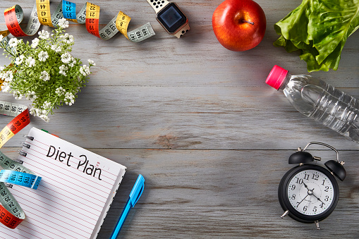 Time to exercise and dieting theme: Top view of Clock, notepad with pen, tape measure, water bottle, apple, towel and dumbbells on a table with copy space