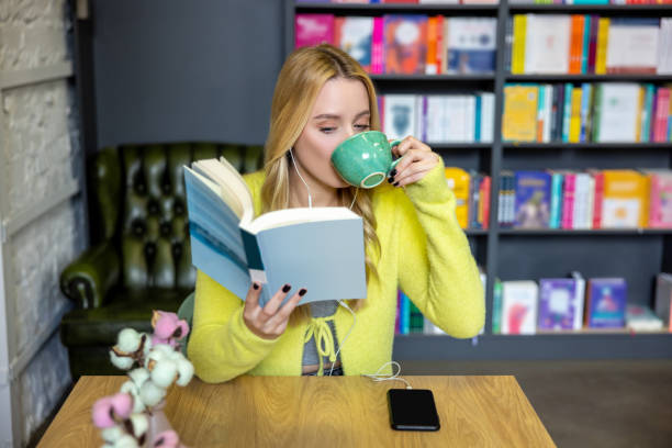 In library. Young blonde woman drinking coffee with a book