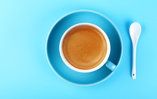Close up one full cup of espresso coffee and saucer with white spoon over pastel blue paper background, elevated top view, directly above