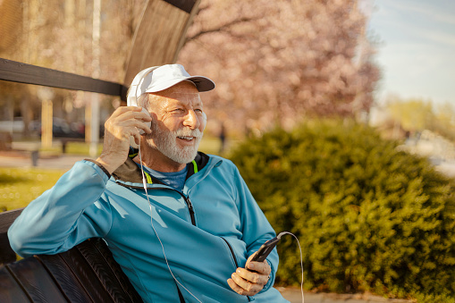 Senior bearded man with smartphone and headphones relax on a park bench. Sports fitness and physical exercises for the elderly