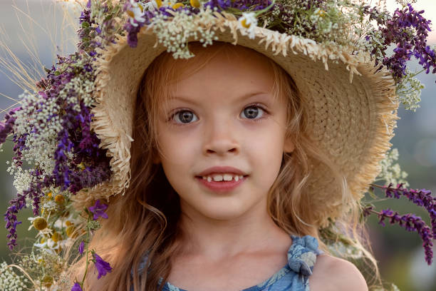 Portrait of cute red-haired little girl in flower crown standing on meadow at sunset. Shooting in the countryside on a summer day