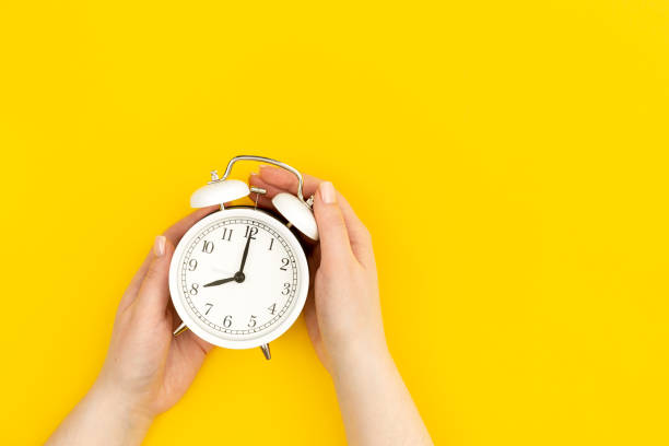 istock White alarm clock in female hands on a yellow background, flat lay, copy space. 1476277499