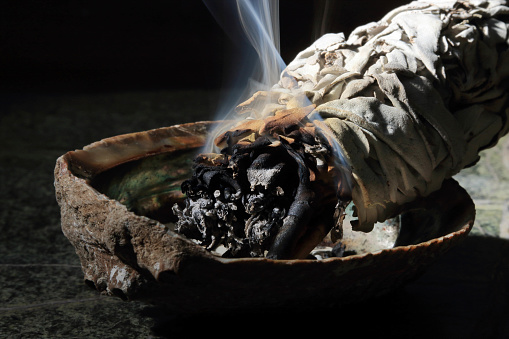 Burning white sage in a seashell on a green granite table with white smoke closeup