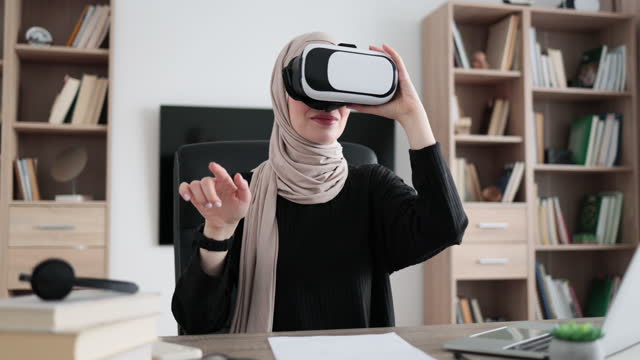 Young attractive muslim woman in hijab sitting at office and using VR headset