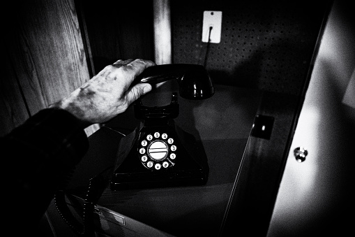 Vintage black and white film noir styled image of a senior man's spooky, wrinkled, gnarly veined hand slowly picking up the receiver on an ancient black landline rotary dial telephone plugged into the background wall with a skinny telephone cable wire.