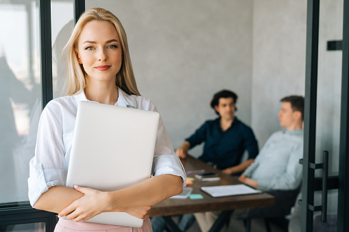 Portrait of attractive female entrepreneur in casual clothes holding laptop in hand standing in office, looking at camera. Startup business team discussing project sitting at desk on background.