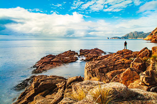 Young man standing by the ocean looking out in golden morning light. Coles Bay, Tasmania, Australia.