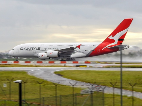 A Qantas Airbus A380-842 plane taking off to the south on a wet runway from Sydney Kingsford-Smith Airport.  The plane, registration VH-OQK, was flying to Singapore as flight number QF1.  In the background is the direction of west and the setting sun. This image was taken from Shep's Mound, a public viewing area off Ross Smith Avenue, Mascot on an overcast and rainy afternoon on 25 March 2023.