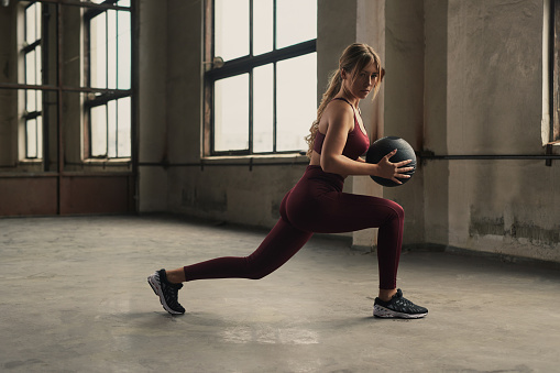 Full body side view of young fit female in sportswear doing lunges with medicine ball in hands during fitness workout in gym