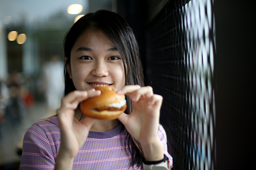 An Asian young woman is enjoying burger in restaurant.