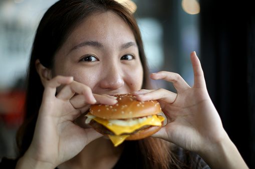 An Asian young woman is enjoying burger in restaurant.