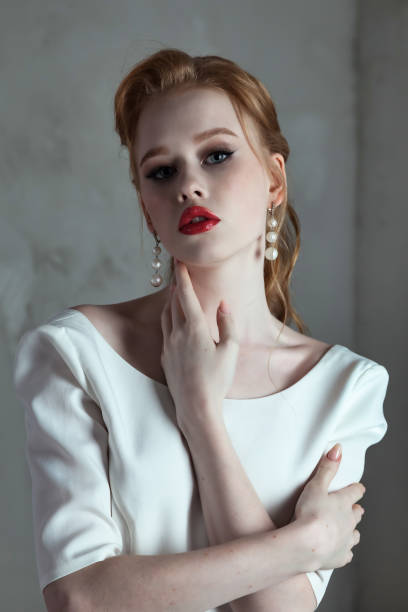 Portrait fashionable bride irish woman with red hair and lips posing at grey background, looking at camera. Stylish lady in white wedding dress in room. Fashion style beauty concept. Copy text space retrato de la novia de moda mujer irlandesa con el pelo rojo y los labios posando sobre fondo gris, mirando a la cámara. elegante dama con vestido de novia blanco en la habitación. concepto de belleza de estilo de moda. copiar espacio de texto