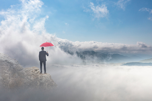 A businessman stands on top of a mountain top as he holds an umbrella as he tries to protect himself from risk.