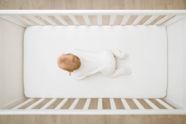 Baby in white bodysuit try crawling on knee and arms on mattress in wooden crib at home room. 5 to 6 months old infant development. Top down view. stock photo