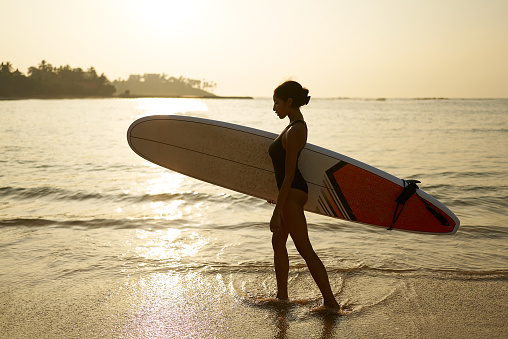 African american woman walking with surfboard on ocean beach. Black female surfer posing with surf board. Pretty multiethnic girl goes on surfing session, enjoys sun at a tropical location at sunrise.