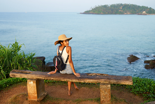Multiethnic woman in straw hat enjoys tropical vacation sitting on rocky cliff with sea view. Black female with backpack sightseeing on scenic location. Girl tanning on island on ocean sunset.