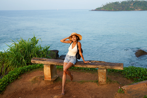 Multiethnic woman in straw hat enjoys tropical vacation sitting on rocky cliff with sea view. Black female with backpack sightseeing on scenic location. Girl tanning on island on ocean sunset.