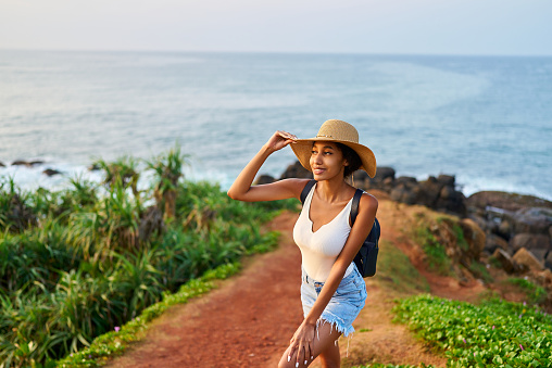 Multiethnic woman in straw hat enjoys tropical vacation travel holiday. Black female with backpack sightseeing on scenic location. Pretty lady standing on island on sunset. Girl in the sun for suntan