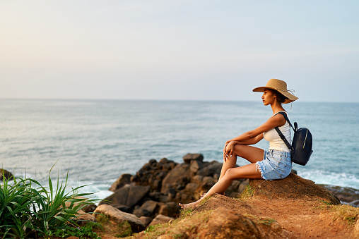 Multiethnic woman in straw hat enjoys tropical vacation sitting on rocky cliff with sea view. Black female with backpack sightseeing on scenic location. Pretty lady tanning on island on ocean sunset.