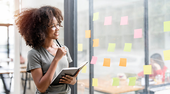 Smiling young black businesswoman holding notebook looking and thinking, standing next to a glass wall