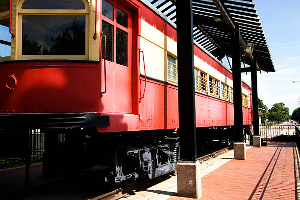 Geary, Blaine County, Oklahoma, USA - May 30, 2024: Vibrant red and yellow Rock Island caboose sits prominently in a grassy area of a small town square in Geary, Oklahoma. A yellow metal playground structure resembling a train stands nearby. Modest brick buildings and cloudy sky form backdrop, capturing essence of rural Americana and railway heritage.