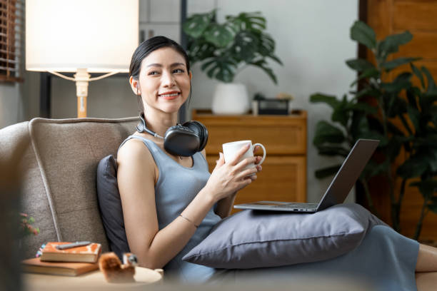 mulher sorridente relaxando em casa, usando laptop, ouvindo música e bebendo café quente. - relaxation success luxury women - fotografias e filmes do acervo