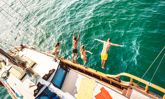 Top view of trendy adventurous friends jumping from sailboat on sea ocean trip - Millennial guys and girls having fun at exclusive boat party - Luxury vacation life style concept on bright filter