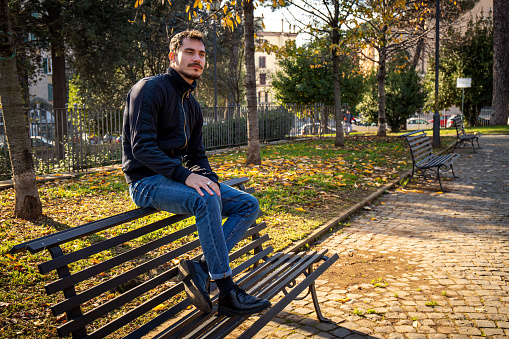 The young man is sitting on the bench in the park in Rome.