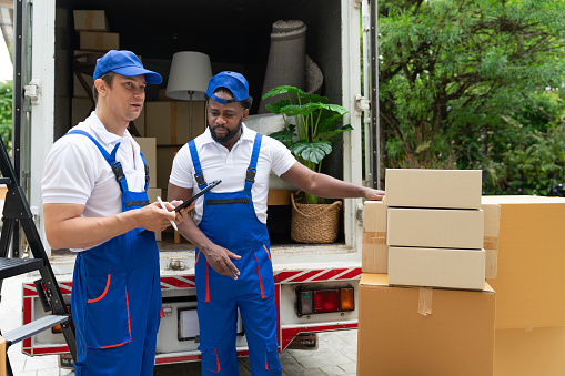 Man mover worker in blue uniform checking lists on clipboard while unloading cardboard boxes from truck.Professional delivery and moving service.