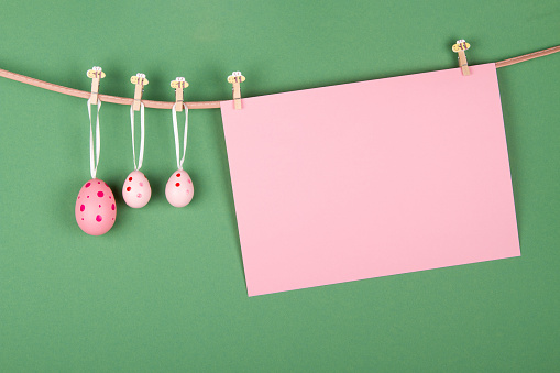 Blank pink sheet of paper and three pink Easter eggs hang at rope on green background. Easter greeting card design with empty space for text