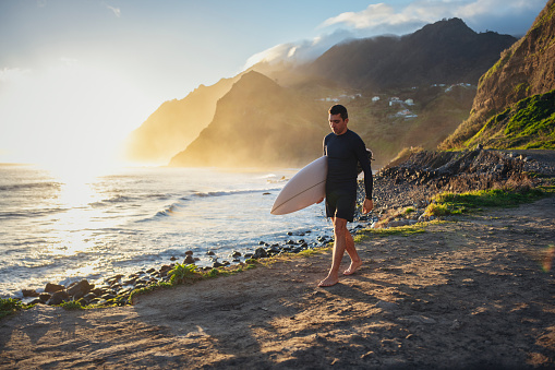 Handsome middle aged surfer walking on a beautiful beach by the ocean at sunrise.