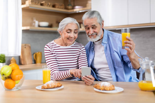 happy senior couple using smartphone in kitchen while having breakfast together - two parent family indoors home interior domestic kitchen imagens e fotografias de stock