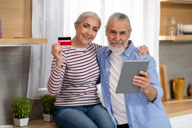 happy elderly couple with digital tablet and credit card posing in kitchen - two parent family indoors home interior domestic kitchen imagens e fotografias de stock
