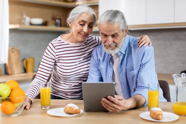 happy senior couple browsing internet on digital tablet during breakfast in kitchen - two parent family indoors home interior domestic kitchen imagens e fotografias de stock