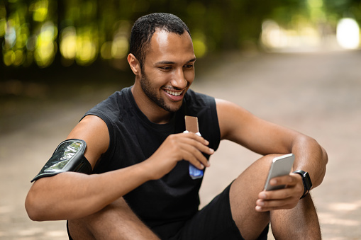 Cheerful handsome black guy sportsman having break while training in public park, sitting on the ground, eating protein bar and using cell phone, watching fitness videos online, copy space