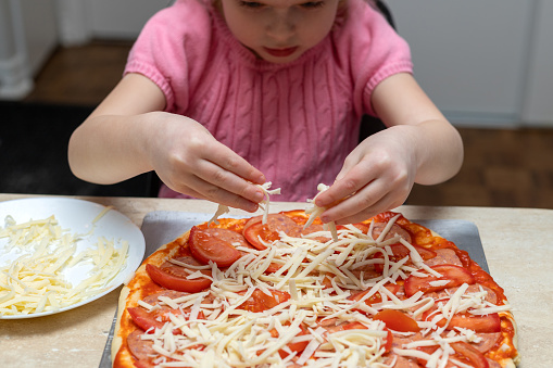 Small child cooking at home sitting at the table. Little girl making pizza in the kitchen. Kid learning to cook