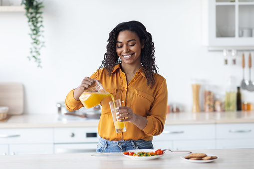 Smiling young black woman in casual pouring fresh orange juice, having healthy vegetable salad, enjoying lunch at home, kitchen interior, copy space. Nutrition, healthy diet concept