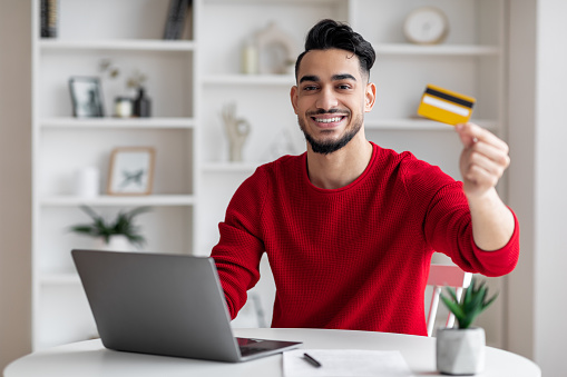 Glad attractive millennial muslim man with beard in red clothes typing on laptop and showing credit card in home office interior. Online shopping, buying, finance and banking during covid-19 pandemic