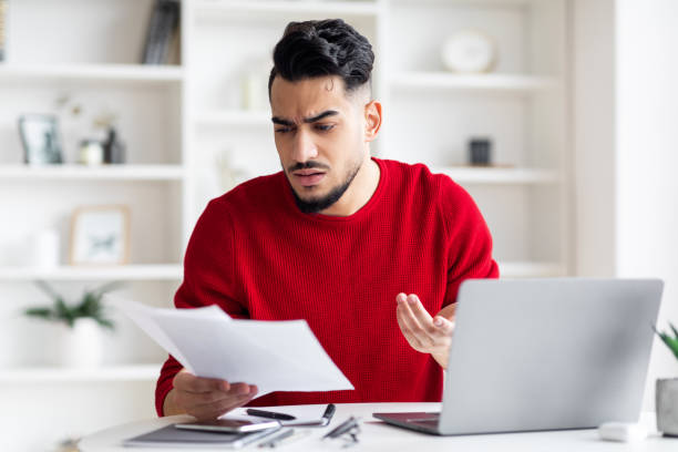 istock Sad worried young arab man with beard works with documents and cant find at workplace with laptop 1476719719