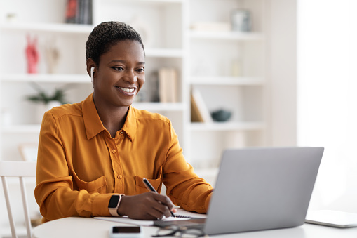 Cheerful short-haired millennial african american woman student having online class at home, sitting at workdesk in front of laptop, using wireless earbuds, taking notes, copy space, e-education