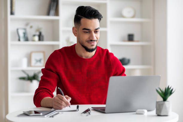 istock Smiling calm confident attractive millennial arab guy with beard in red clothes makes notes, works on laptop 1476720870