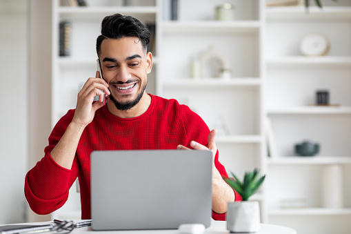 Smiling attractive millennial arab male with beard in red clothes speaks by phone and looks at laptop in home office interior. Work with client remotely, business and tech due covid-19 quarantine