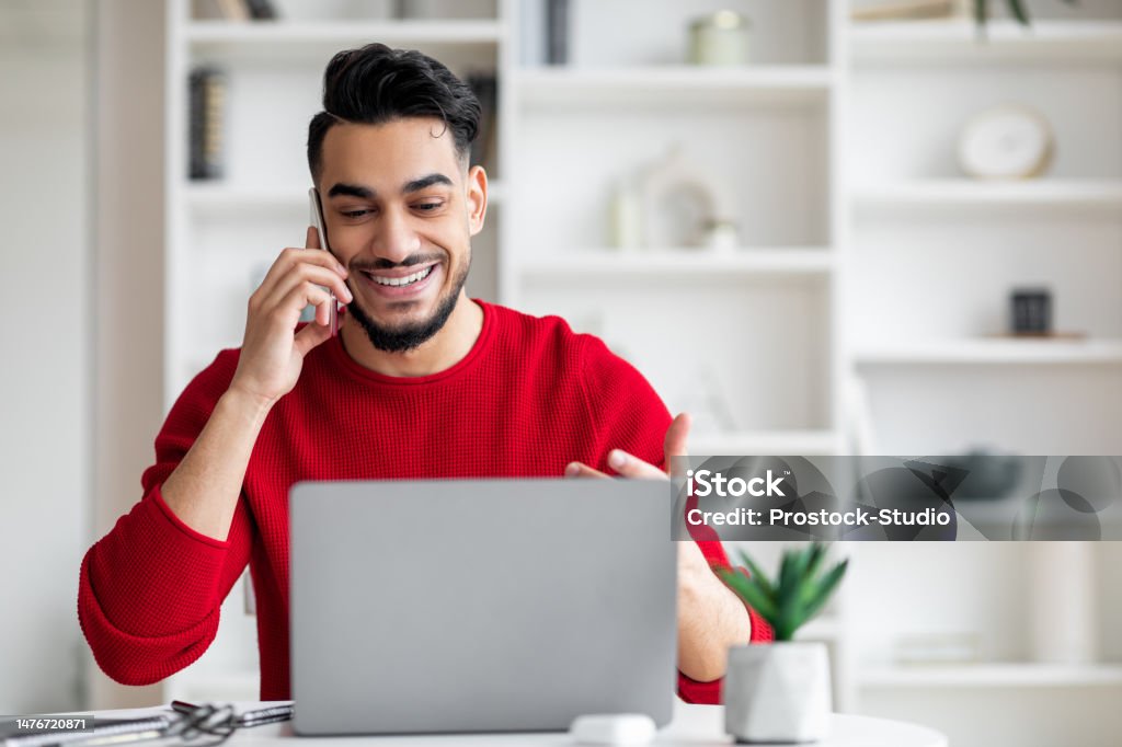 Smiling attractive millennial arab male with beard in red clothes speaks by phone and looks at laptop Smiling attractive millennial arab male with beard in red clothes speaks by phone and looks at laptop in home office interior. Work with client remotely, business and tech due covid-19 quarantine People Stock Photo