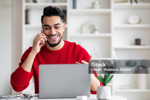 Smiling Attractive Millennial Arab Male With Beard In Red Clothes Speaks By Phone And Looks At Laptop Stock Photo - Download Image Now