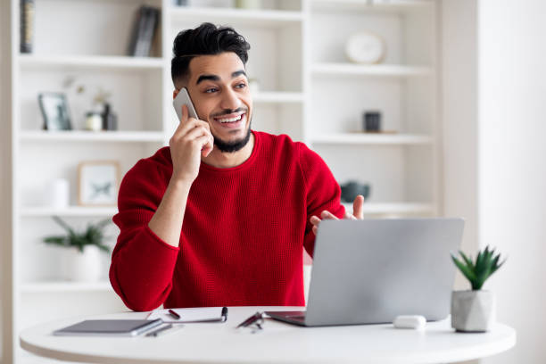 istock Glad young arabic male with beard speaks on phone with client at workplace with laptop in home office interior 1476720877